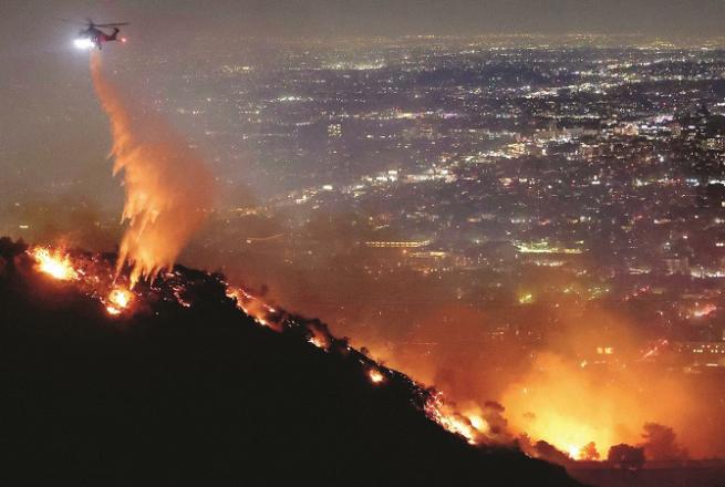 A portion of the iconic Hollywood Hills can be seen engulfed in flames late Tuesday night, with the Los Angeles suburbs glowing in the background and a firefighter helicopter spraying water above. Photo: INN